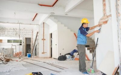 Ouvrier en tenue de travail et casque jaune effectuant des travaux de rénovation sur un mur intérieur.